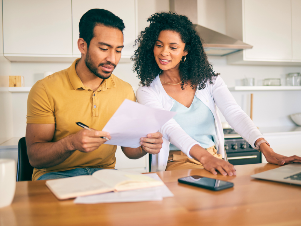 a man with dark hair and medium brown skin sits at a kitchen table with a woman with dark curly hair and medium brown skin reviewing the questions to ask a mortgage lender before meeting to buy a home